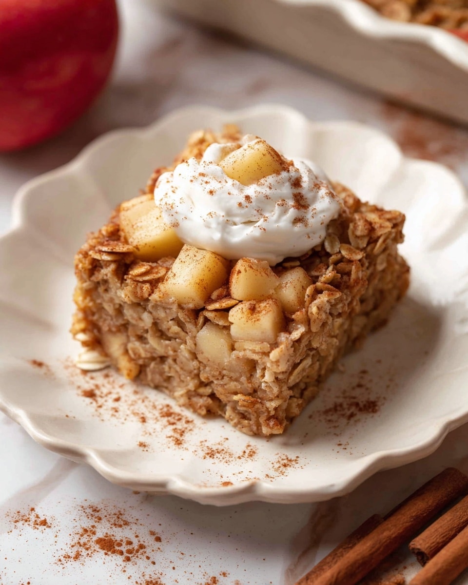 A square piece of oatmeal apple bake sits in the center of a white scalloped plate, showing two layers: the bottom dense baked oatmeal layer with visible bits of apple inside, and the top layer which is a mix of oats and small apple chunks with a slightly rough texture. On top, there is a dollop of white cream sprinkled lightly with cinnamon powder. Around the plate, faint cinnamon powder is scattered. The background has a white marbled texture with part of a red apple, cinnamon sticks, and a dish partially visible out of focus. photo taken with an iphone --ar 4:5 --v 7