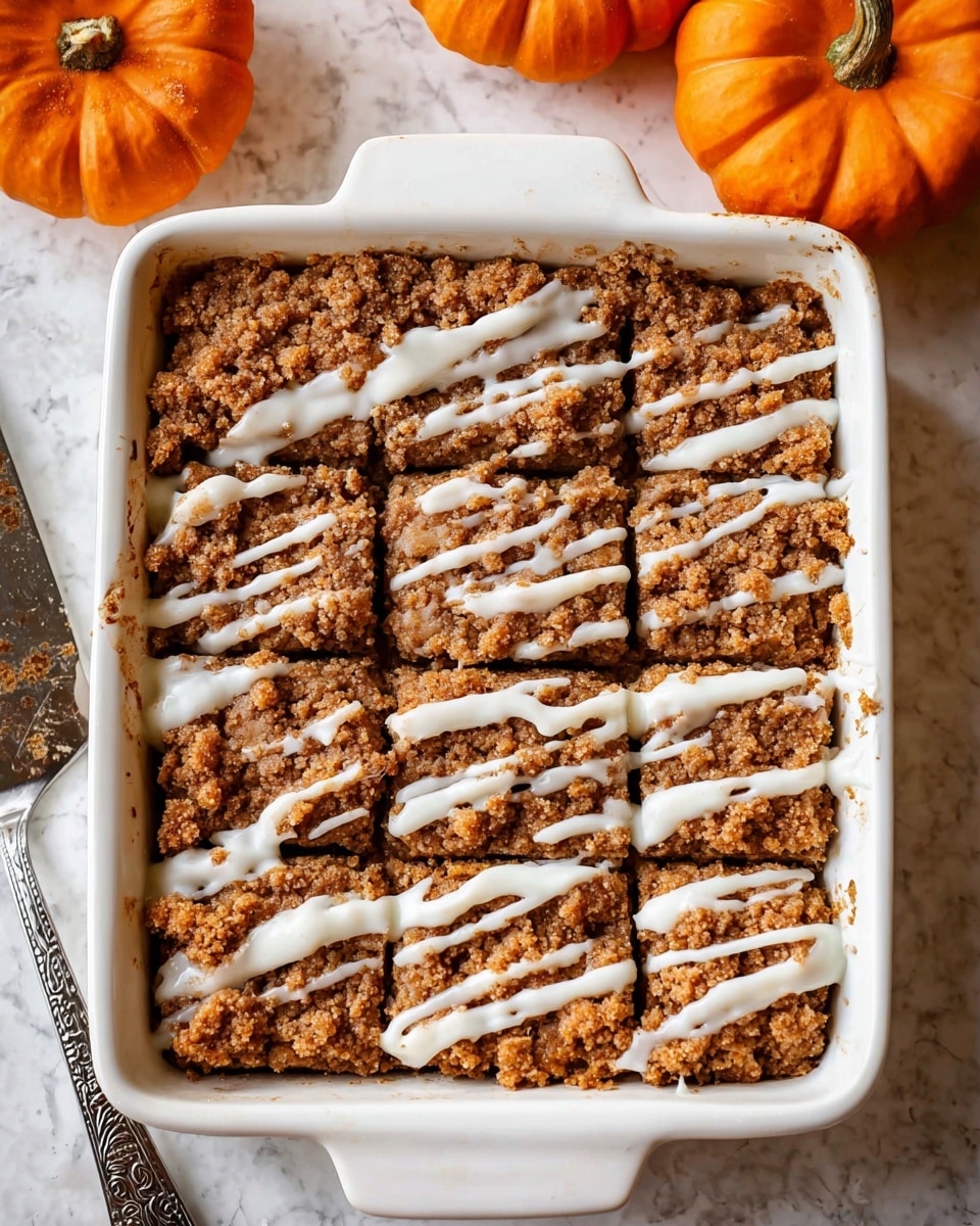 A white baking dish filled with a dessert cut into nine squares, topped with a thick layer of crumbly brown streusel that looks crunchy and textured. Over the streusel, white icing is drizzled lightly in irregular stripes across the top. The dish is placed on a white marbled surface with two small orange pumpkins in the background, adding a warm and cozy feeling. A silver serving spatula with a detailed handle rests partially under the dish on the left side. The top crust layer is uneven with varying sizes of crumbs visible, suggesting a homemade, rustic style photo taken with an iphone --ar 4:5 --v 7